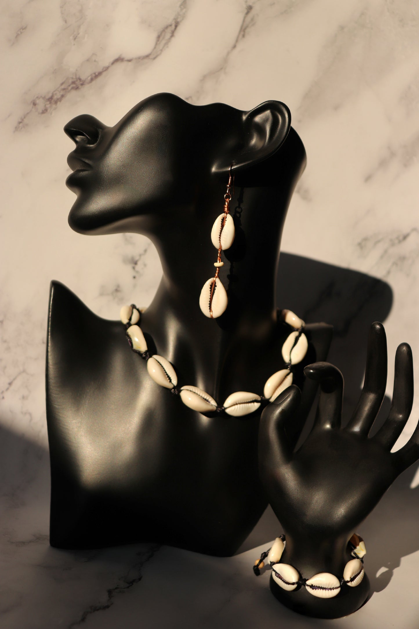 A photo of one handcrafted cowrie shell earring in a mannequin's ear, a bracelet on a mannequin's wrist, and one necklace around a mannequin's neck